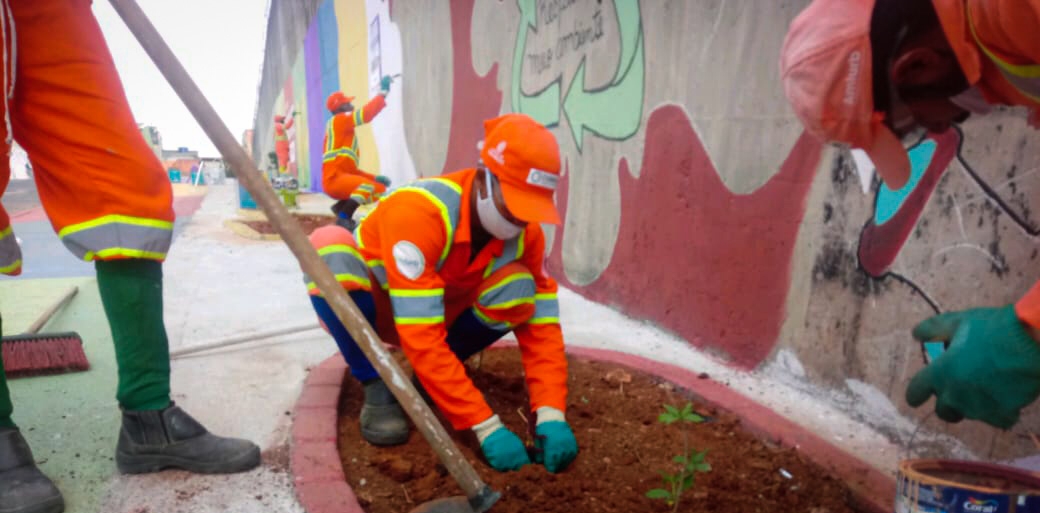 Trabalhadores da limpeza urbana, vestidos com uniformes laranja com faixas refletivas, realizam a revitalização de um espaço público. No primeiro plano, um dos trabalhadores está agachado, plantando uma muda em um pequeno canteiro com solo recém-revolvido. Outro funcionário segura uma ferramenta enquanto auxilia na preparação do terreno. Ao fundo, mais agentes participam da ação, incluindo um que está pintando um muro com grafites coloridos e mensagens escritas, transformando a área em um ambiente mais agradável e sustentável.